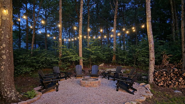 The stone fire pit at dusk, string lights overhead, the woods close on every side
